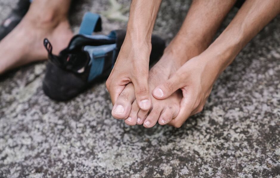 Osoba trzymająca dłońmi stopę obok butów wspinaczkowych na skalistym podłożu.