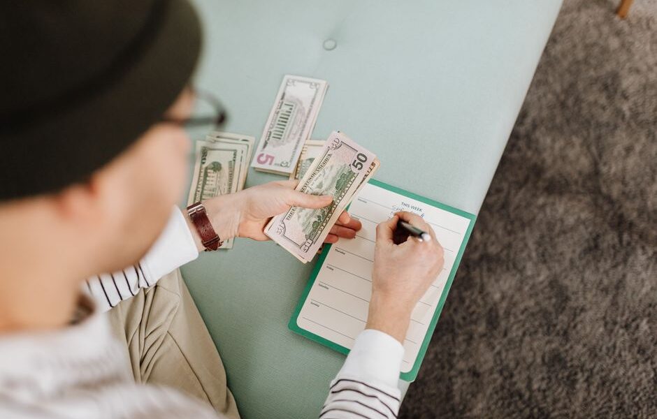 Osoba trzymająca plik dolarów i zapisująca wydatki w notatniku